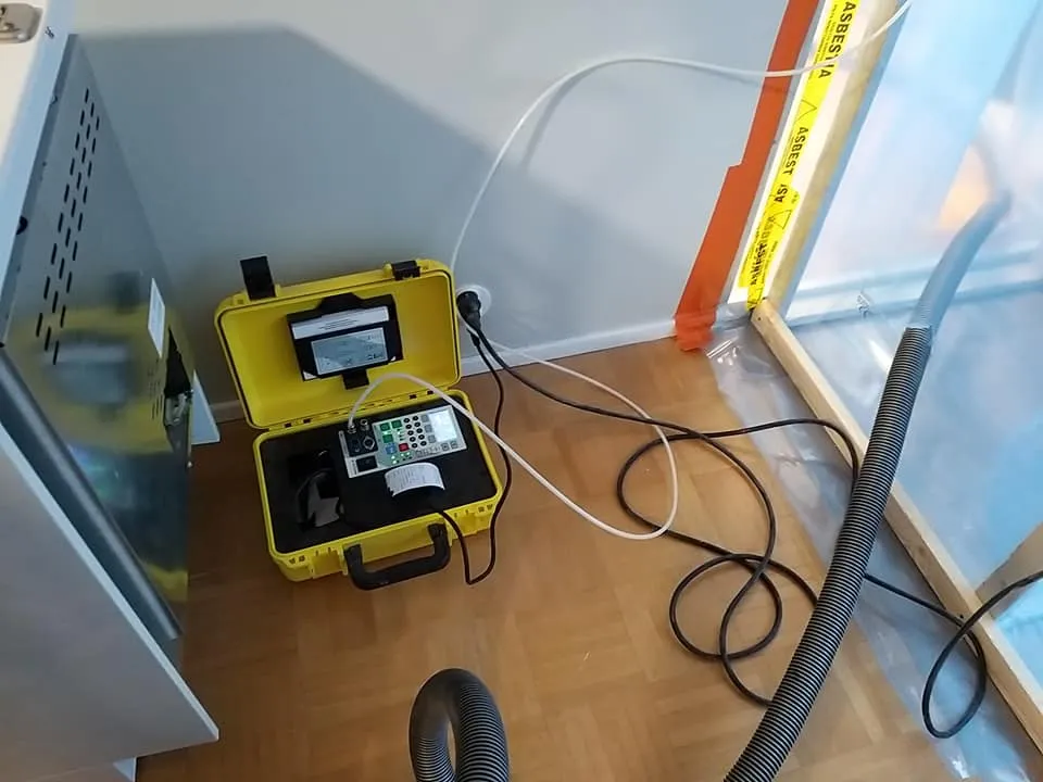 Kuvassa näkyy keltainen laukku, jossa on mittauslaite, sekä ilmastointiputki ja muovilla suojattu alue, jossa on varoitusteippi "ASBESTI" kirjoitettuna.