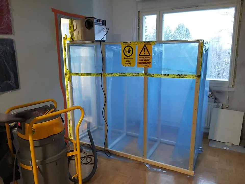 Huoneessa on asbestinpoistoon tarkoitettu suojatila, jossa on muoviseinät ja varoitusteippi. Ikkuna näkyy taustalla.