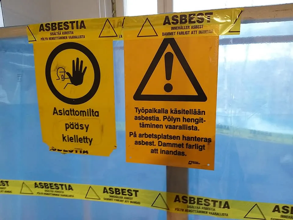 Kaksi varoituskylttiä, joissa lukee "Asbestia" suomeksi ja ruotsiksi, sekä varoitusnauha, joka ilmoittaa asbestista, valaistus on kirkas.