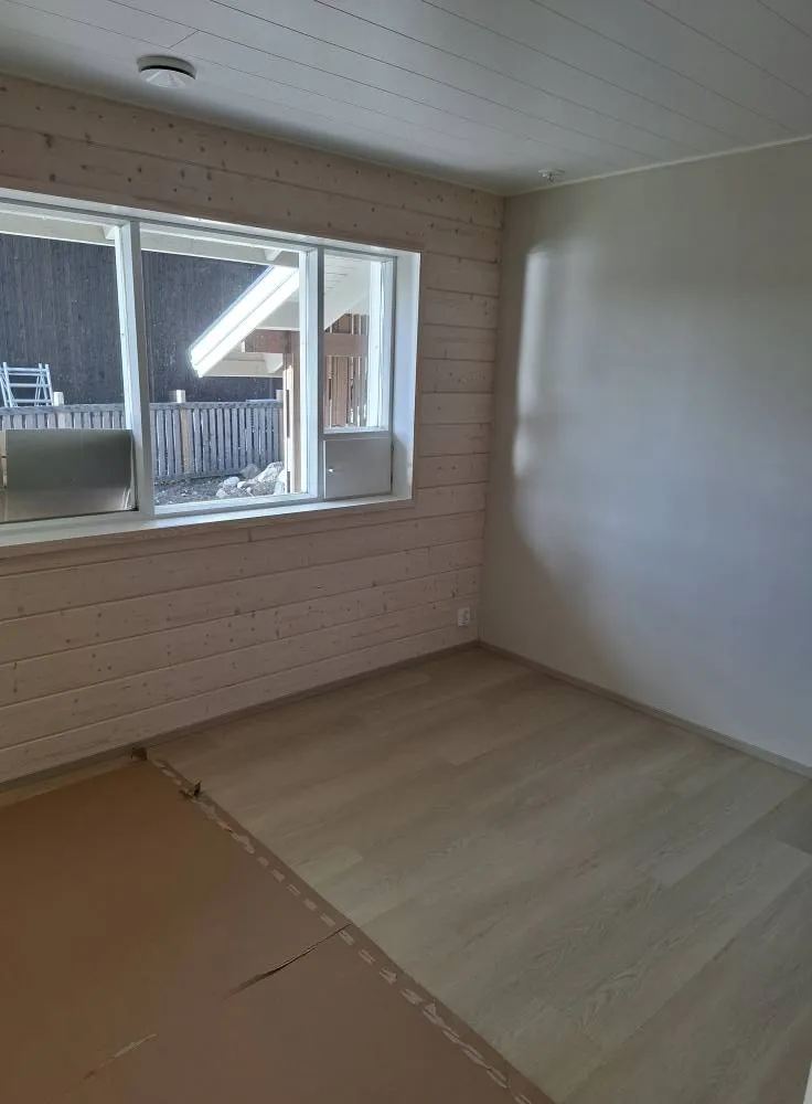 Huoneessa on ikkuna, puupanelointi ja vaalea laminaattilattia, osittain peitettynä ruskealla pahvilla.