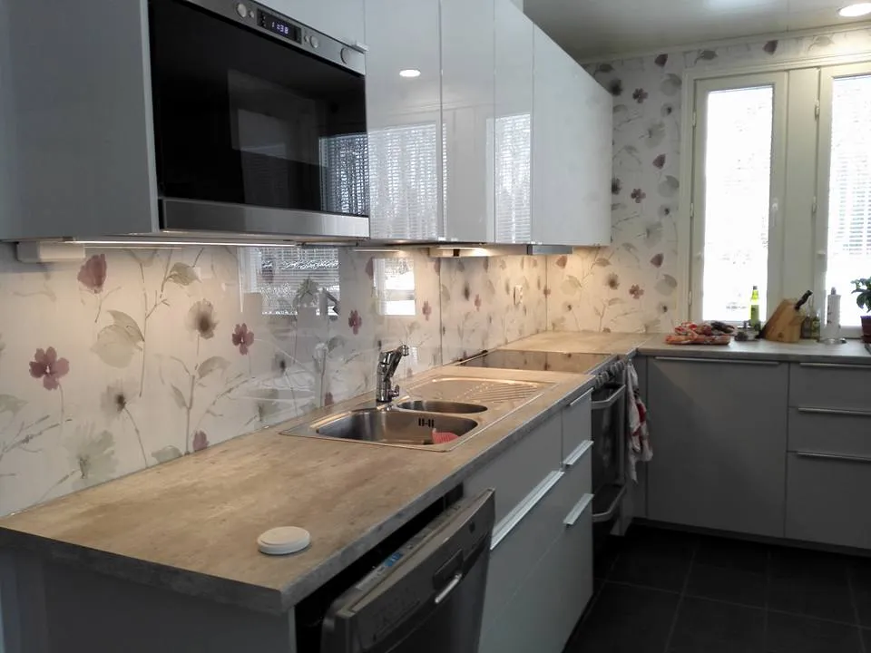 Keittiön sisustus, jossa on valkoiset kaapit, kukkatapetti ja harmaa työtaso. Valoisa tila, jossa on ikkuna.