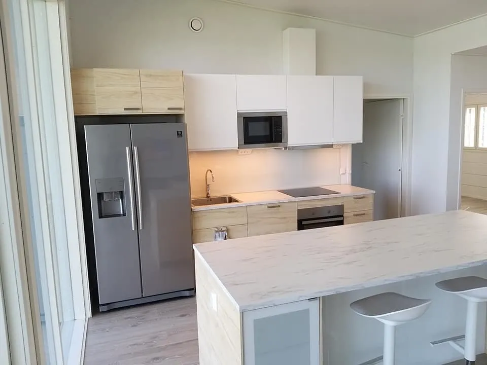 Vaaleassa keittiössä on harmaa jääkaappi, valkoiset ja puunväriset kaapit, marmorinen saareke ja kaksi baarijakkaraa.