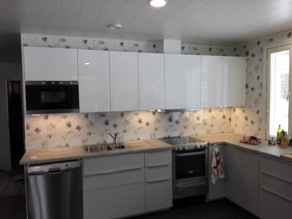 Keittiön sisustus, valkoiset kaapit, kukkatapetti ja ruostumattomasta teräksestä valmistetut laitteet valaistussa tilassa.