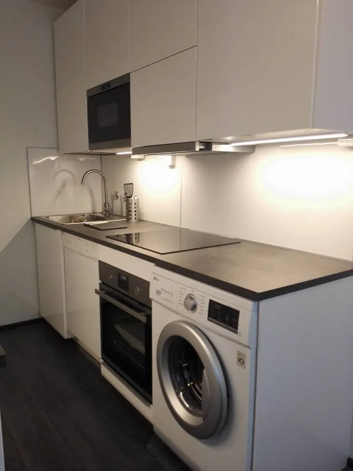 Moderni keittiö, jossa valkoiset kaapit, harmaa työtaso, pesukone ja uuni. Valoisa tila.