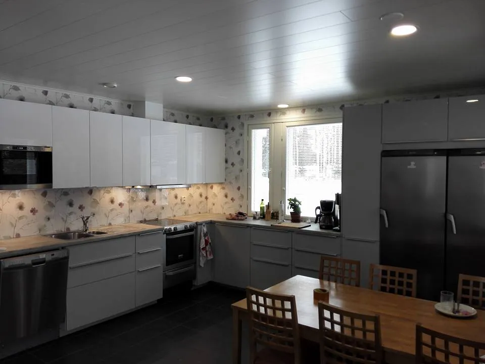 Keittiön sisustus valkoisilla kaapeilla, puupöydällä ja ruokatuoleilla, jossa on kukkatapetti ja kirkas valaistus.