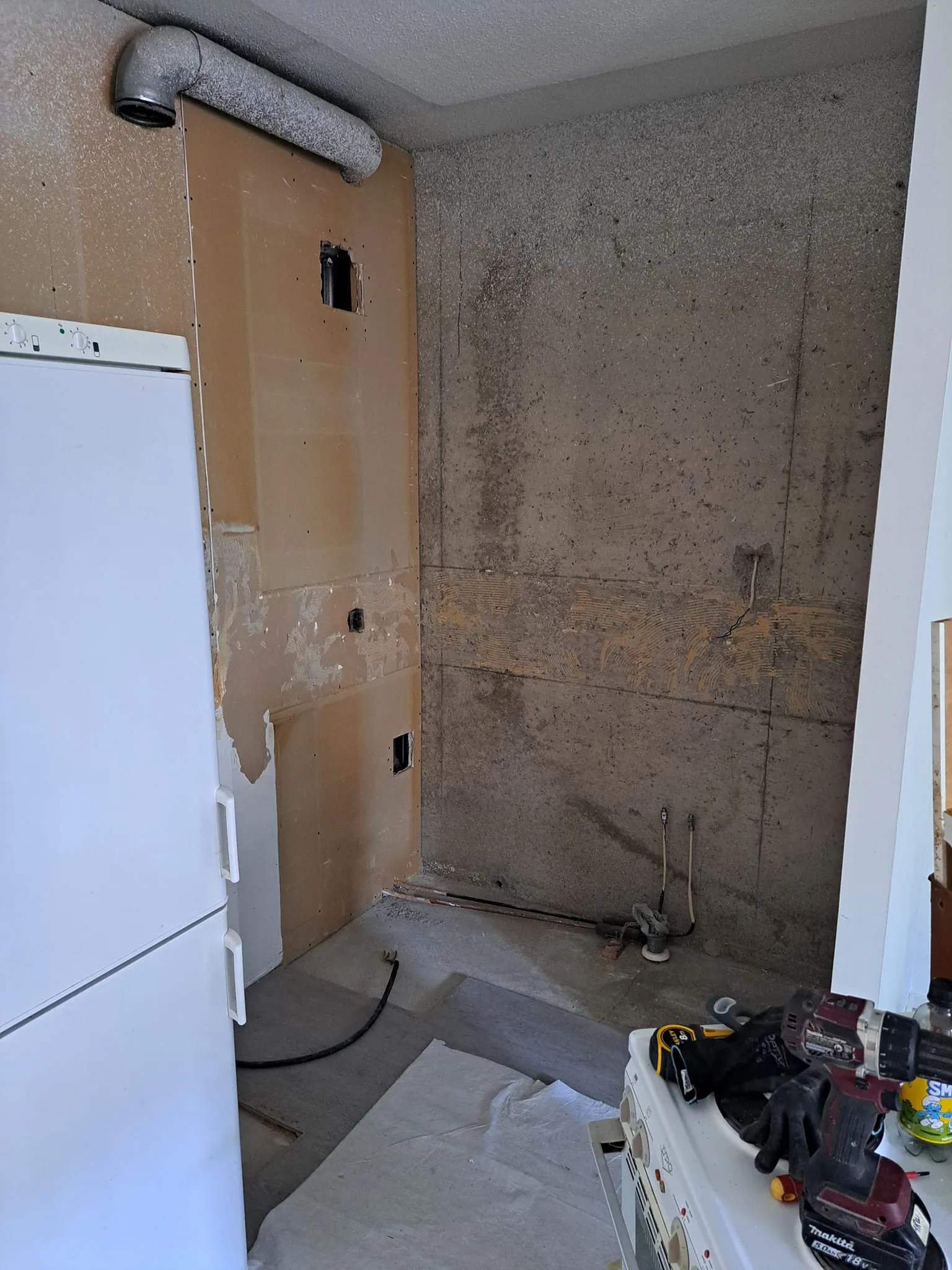 Keskeneräinen keittiö, jossa on valkoinen jääkaappi, paljas seinä ja työkaluja lieden päällä.