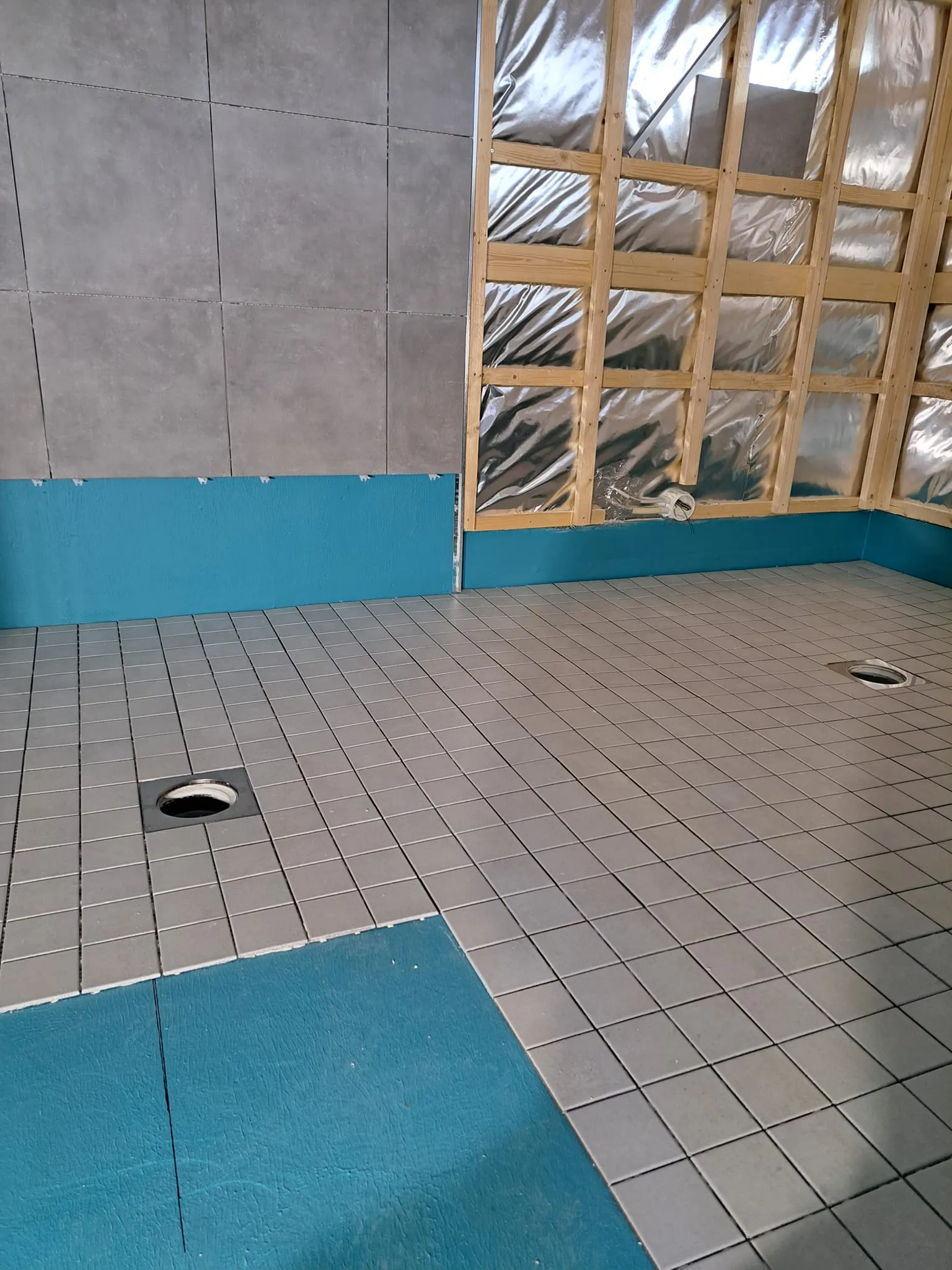 Keskeneräinen kylpyhuone, jossa on harmaat laatat, sininen pohja ja puinen kehys seinällä.