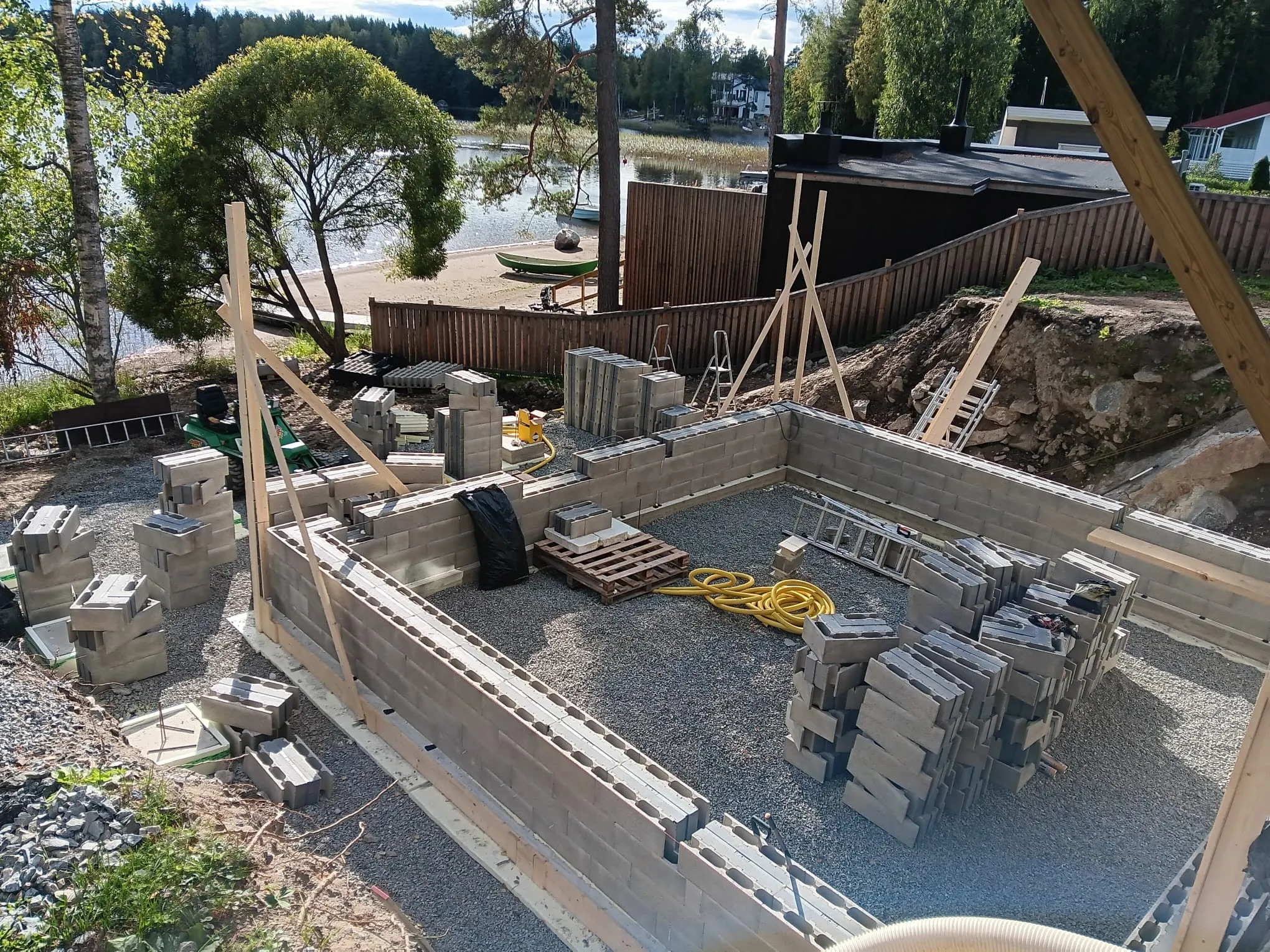 Rakenteilla oleva kivijalka, jossa harmaita harkkoja, keltaisia letkuja ja puutavaraa, valoisassa ulkotilassa.