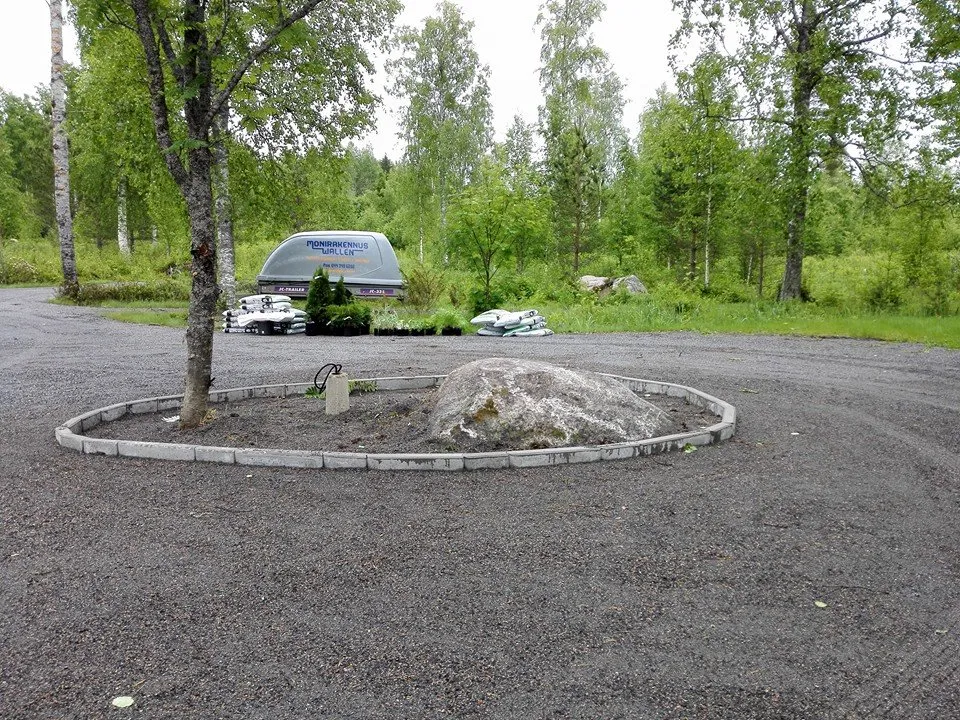 Ulkona on kivikkopiha, jossa on pyöreä kivireunus, suuri kivi, puu ja taustalla metsää sekä peräkärry.