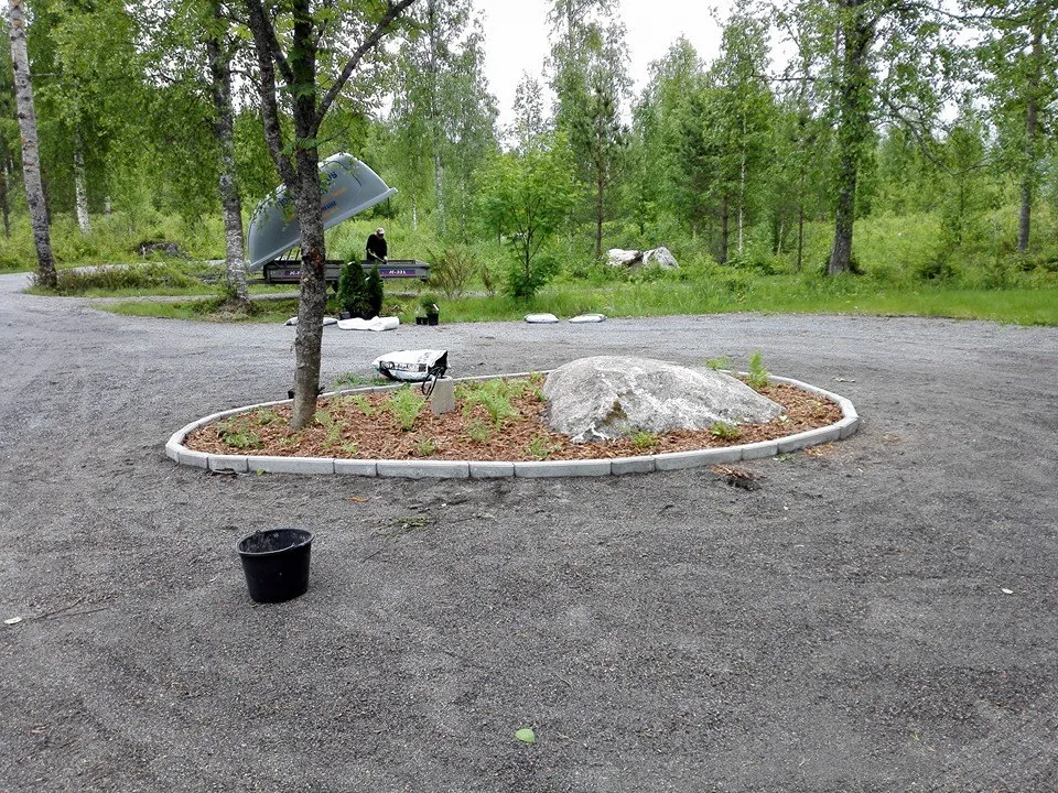 Ulkona on kivikkopuutarha, jossa on suuri kivi ja puu, sekä musta ämpäri ja peräkärry taustalla.