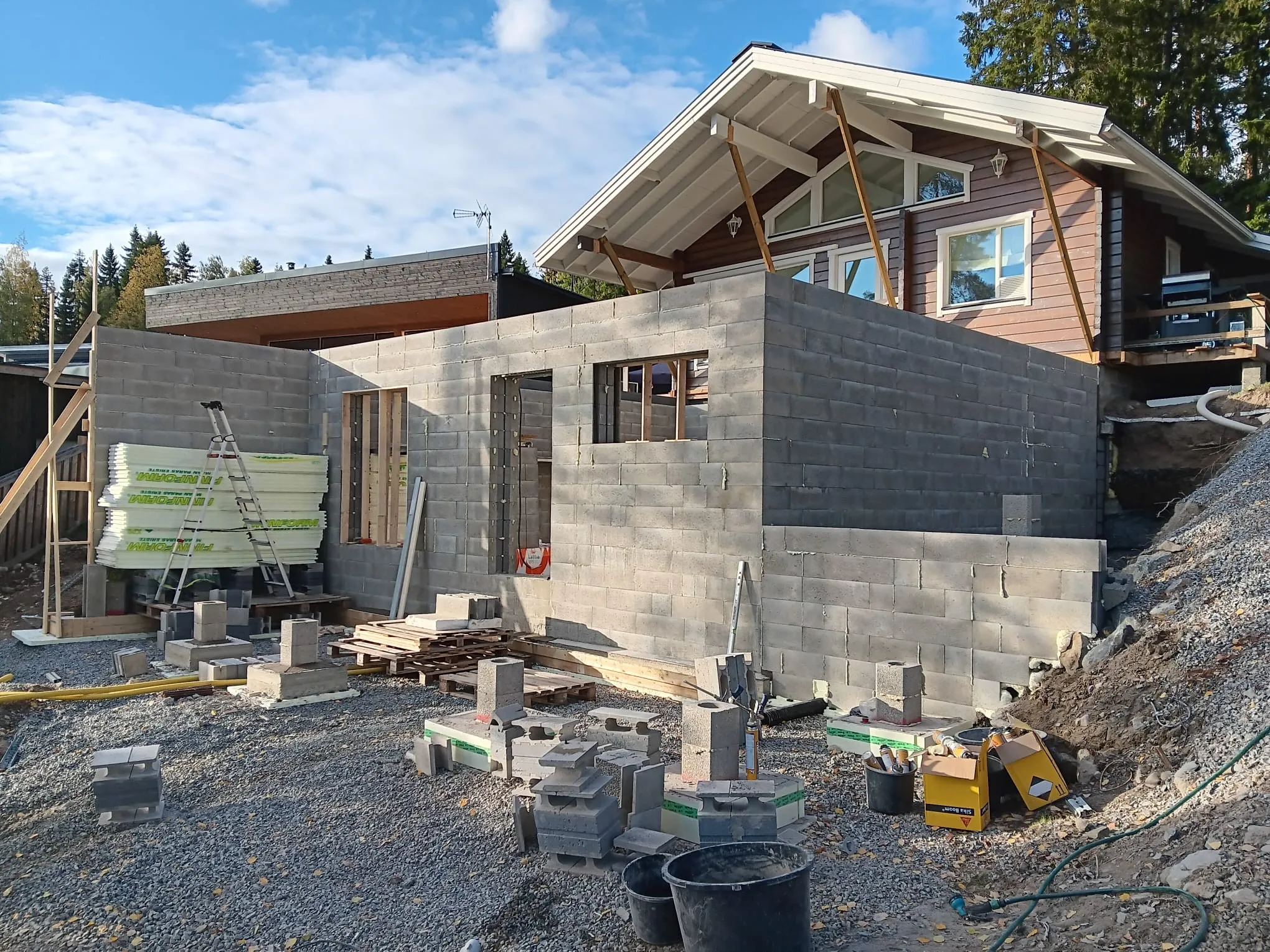 Rakenteilla oleva harmaa kivitalo, jossa on puinen yläkerta ja rakennustarvikkeita kivipohjan ympärillä, valoisassa säässä.