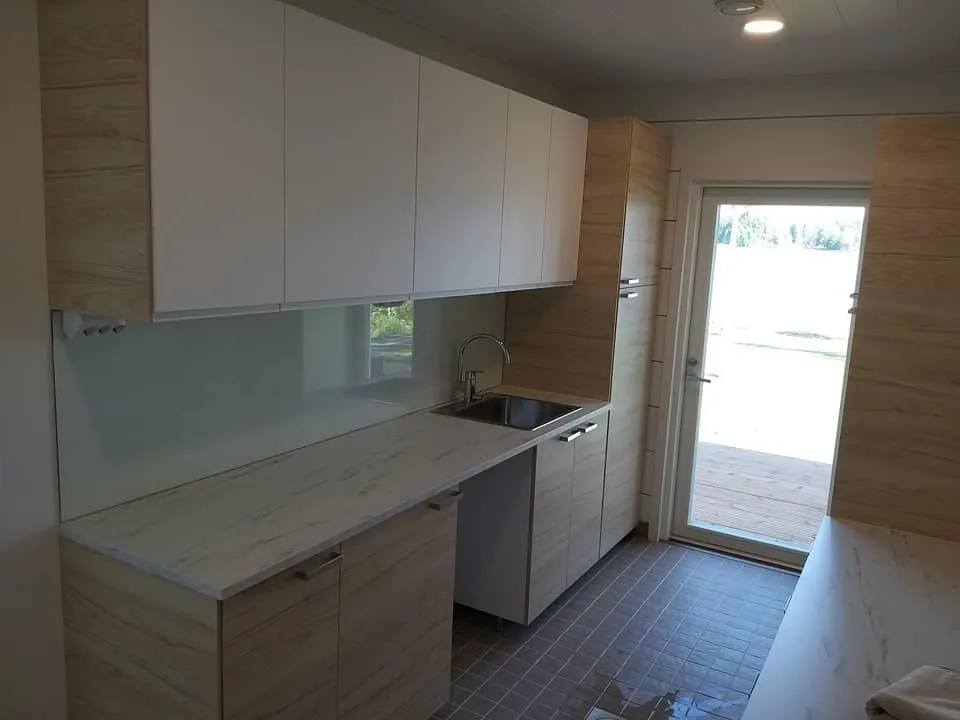 Valoisa kodinhoitohuone, jossa on valkoiset yläkaapit, puukuvioiset alakaapit ja työtaso, sekä teräksinen pesuallas. Lattialla on harmaa laatoitus ja huoneesta on pääsy ulos lasiovesta.