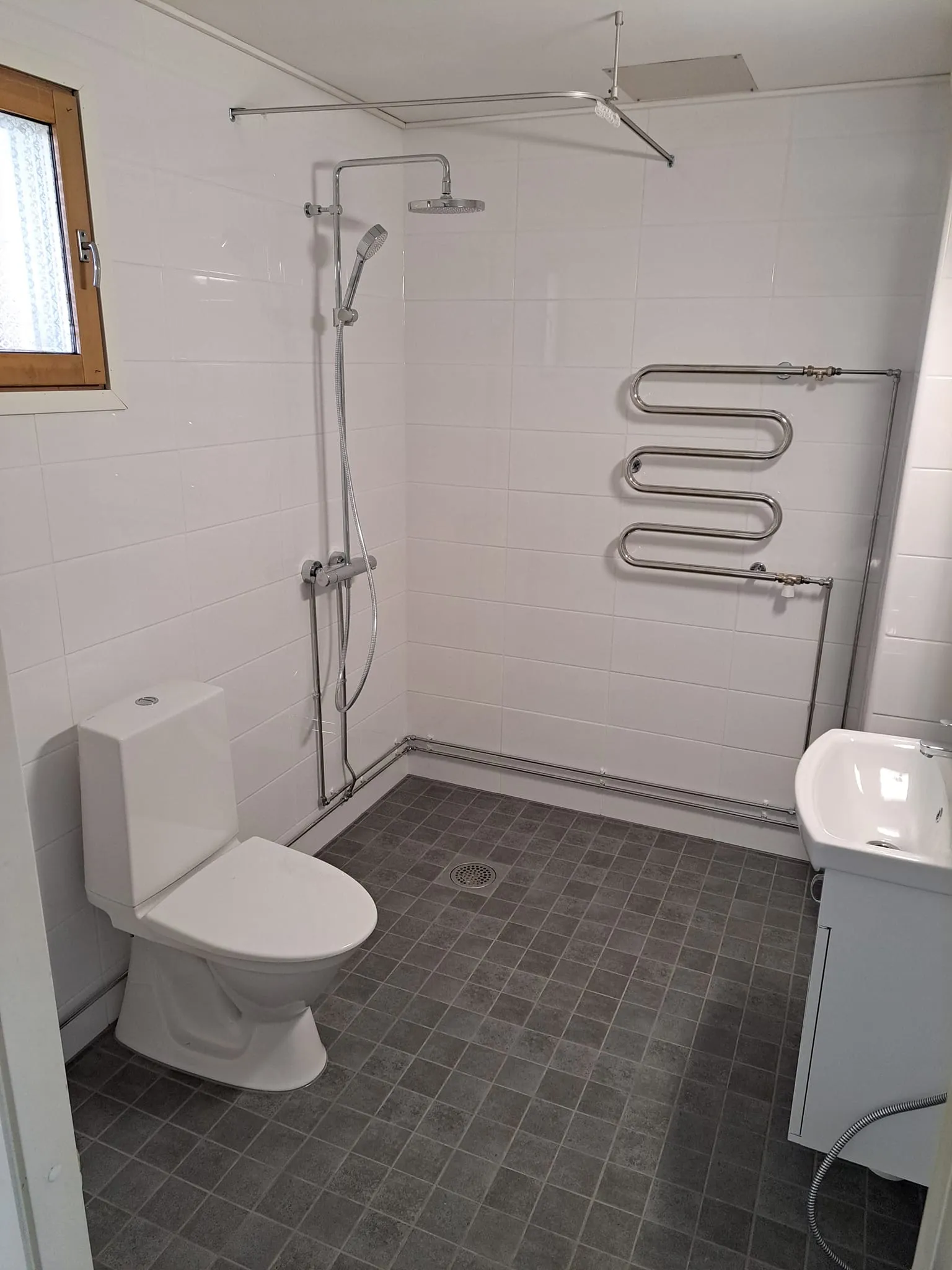 Kylpyhuoneessa on valkoiset seinälaatat, harmaa laattalattia, valkoinen wc-istuin ja pesuallas, sekä metallinen pyyhekuivain ja suihku. Ikkuna on puukehyksinen.