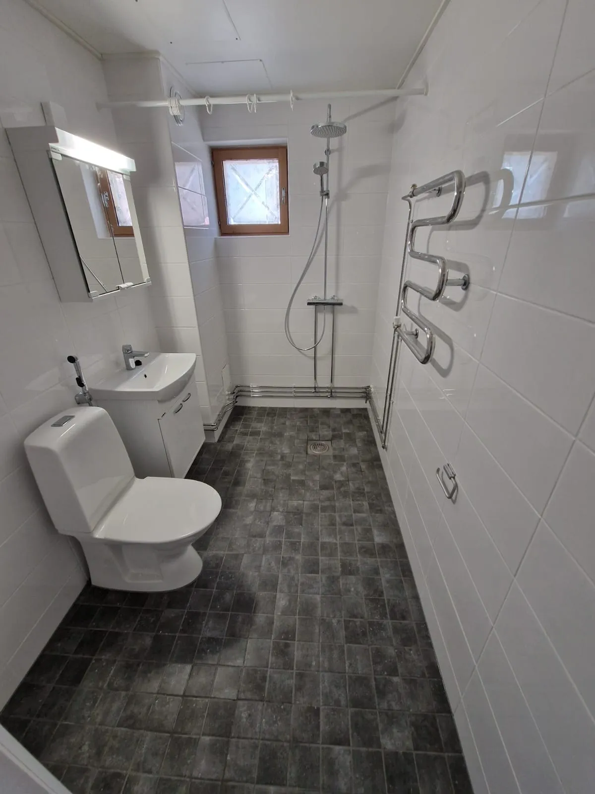 Valoisa kylpyhuone, jossa on harmaat lattialaatat, valkoinen wc-istuin, pesuallas ja suihku. Pyyhekuivain on seinällä.