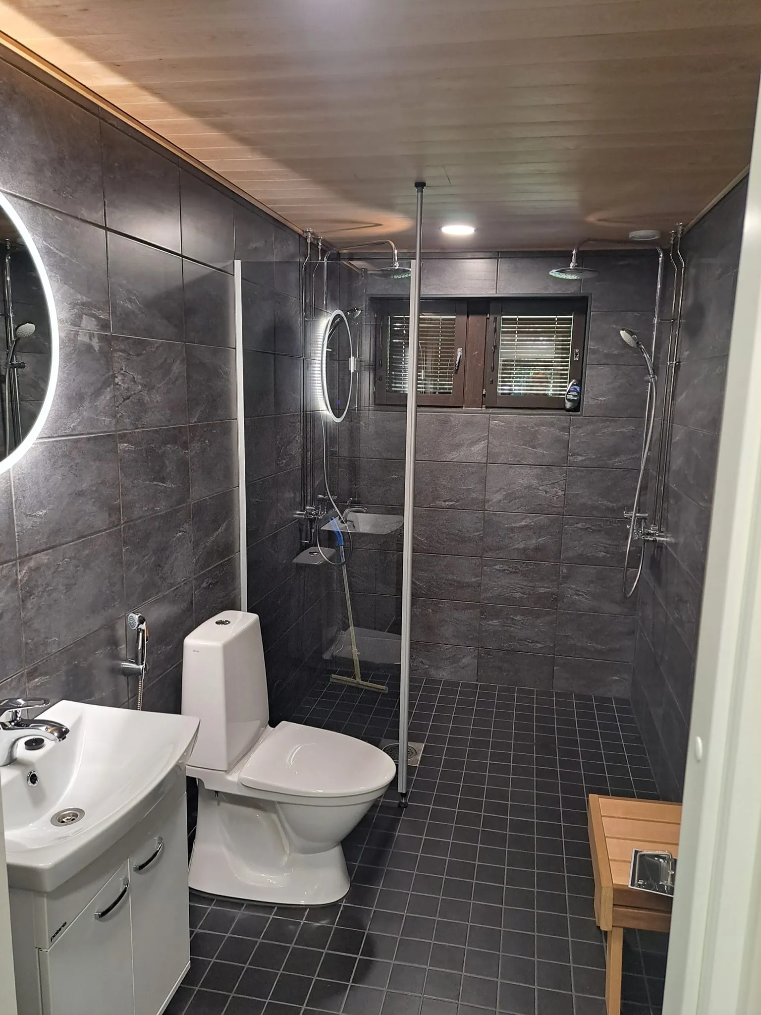 Kylpyhuoneessa on harmaat seinälaatat, puinen katto, valkoinen pesuallas ja wc-istuin sekä suihkukaappi.