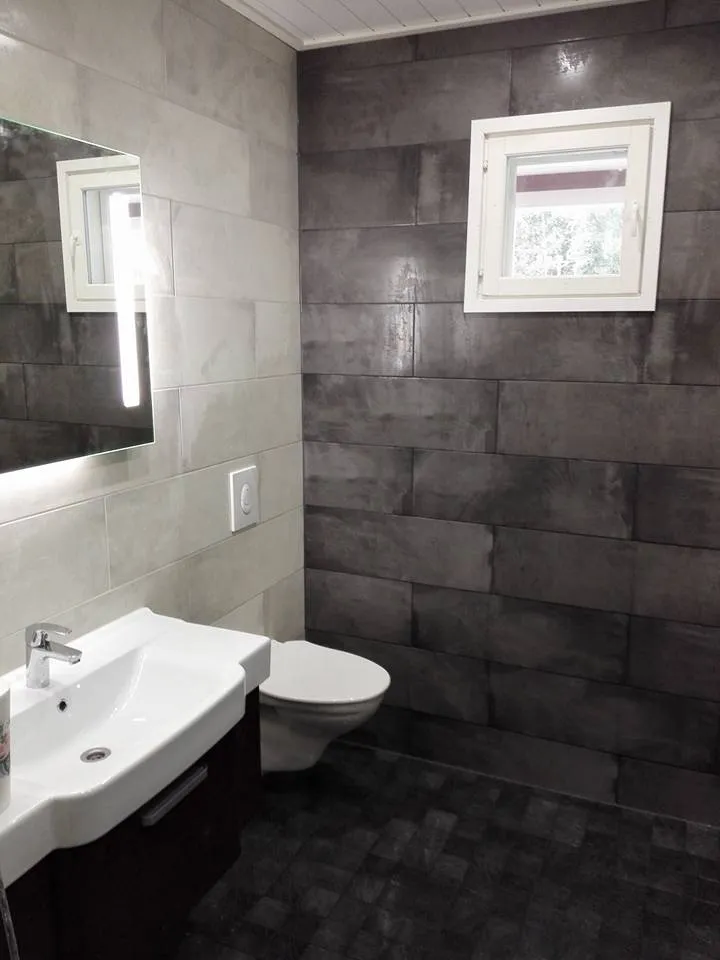 Moderni kylpyhuone, jossa on harmaat laatat, valkoinen pesuallas ja peili valolla.