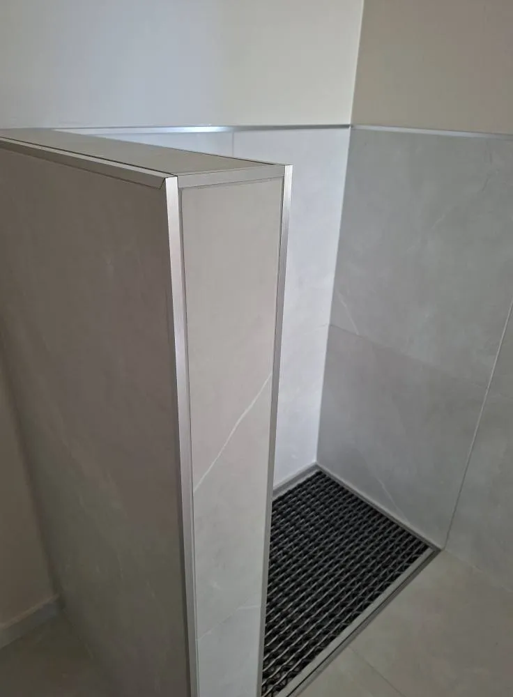 Kylpyhuoneen kulmaus, jossa on harmaa seinä, metallinen ritilä ja alumiinikehykset. Valo on kirkas ja tasainen.