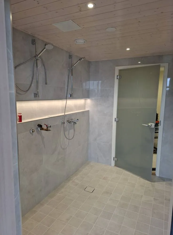 Kylpyhuoneen sisätila, jossa on kaksi suihkupäätä, laatoitettu lattia ja seinät sekä himmeä lasiovi. Puinen katto ja valaistus luovat modernin tunnelman.