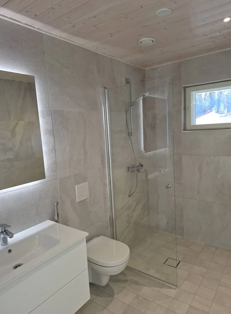 Kylpyhuoneessa on valkoinen pesuallas, peili, wc-istuin ja suihkukaappi, jossa on harmaa laatoitus ja puinen katto.