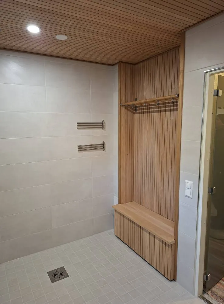 Kylpyhuoneen sisustus, jossa on puinen katto, valkoiset seinälaatat, pyyhetelineet ja puinen vaatekaappi, jossa on penkki ja vaatetanko.