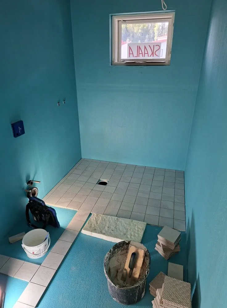 Valoisa kylpyhuone rakennusvaiheessa, jossa turkoosit seinät, lattialaatat osittain asennettu, laastipalju ja ikkuna.