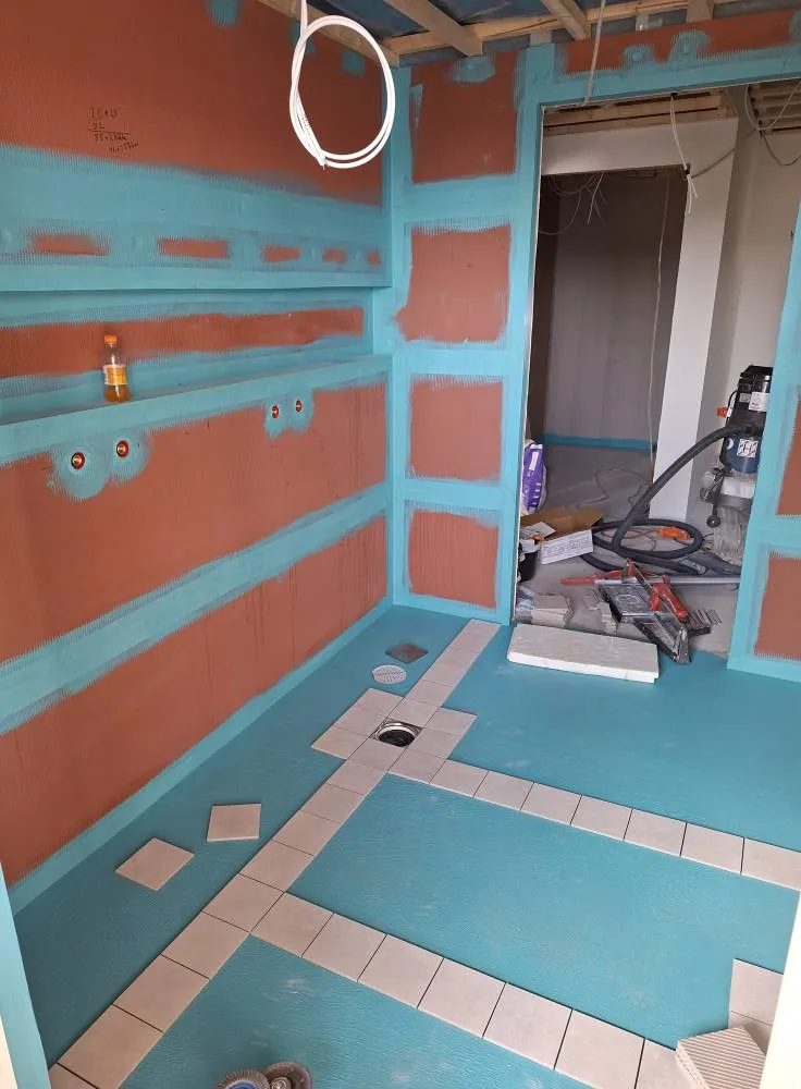 Rakenteilla oleva kylpyhuone, jossa on sininen vedeneristys, ruskeat seinät ja lattialaatat osittain asennettu.