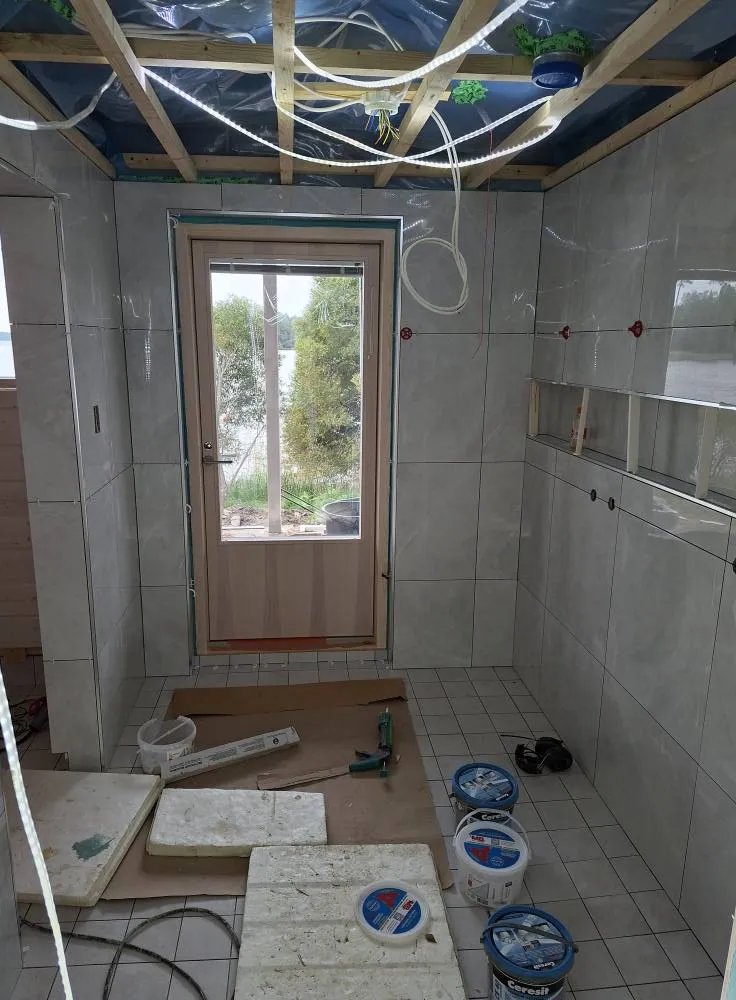 Keskeneräinen kylpyhuone, jossa on laatoitetut seinät, puinen ovi, työkalut ja Cerestit-ämpärit lattialla, sekä paljas katto.