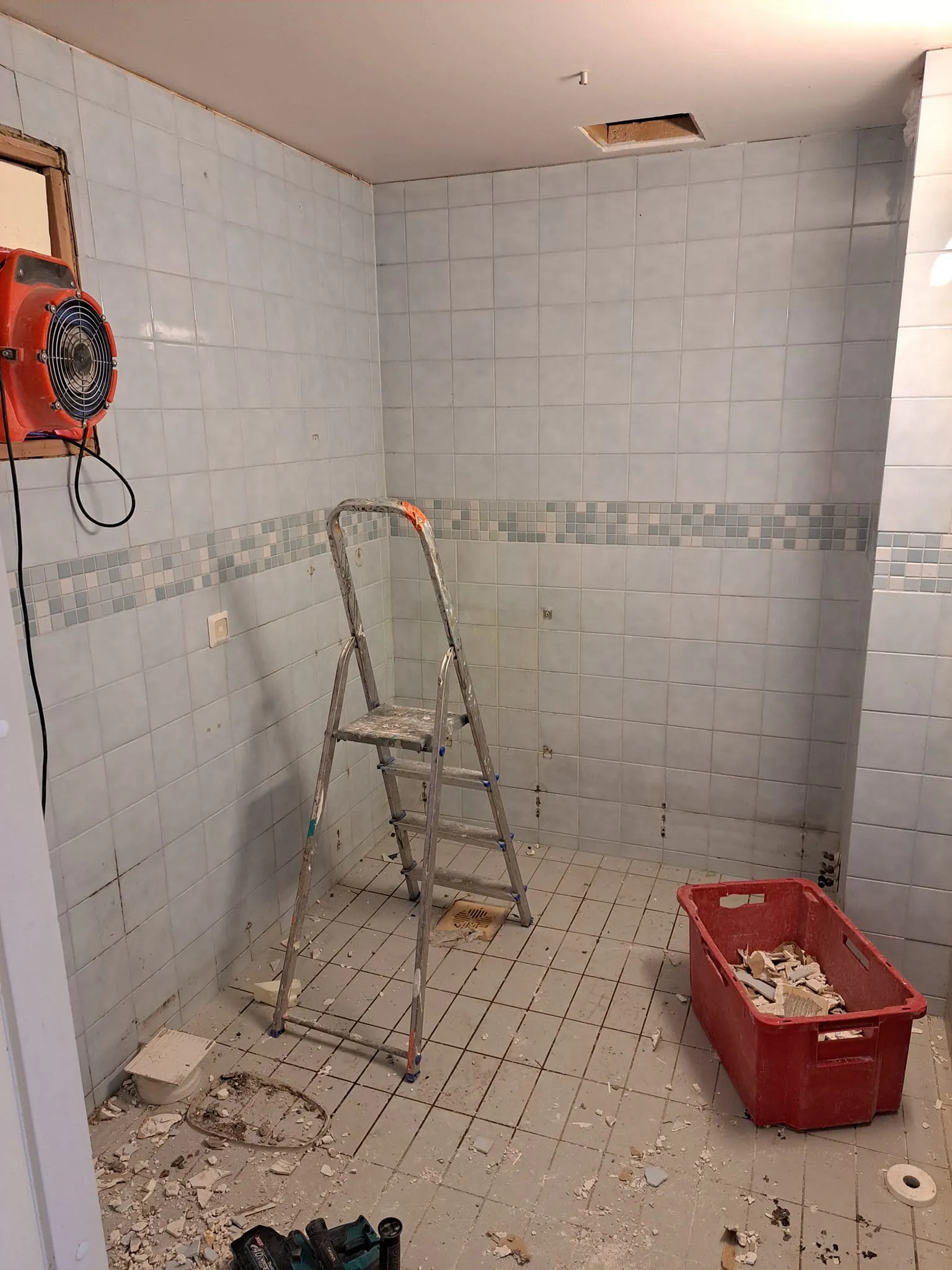 Remontissa oleva kylpyhuone, jossa on tikkaat, punainen laatikko ja oranssi tuuletin. Laatoitetut seinät ja lattia, valaistus on kirkas.