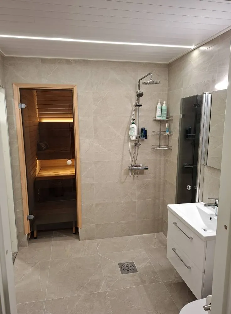 Kylpyhuoneen sisustus, jossa on sauna, suihku, pesuallas ja harmaat laatat. Valoisa tila, jossa on puuta ja metallia.