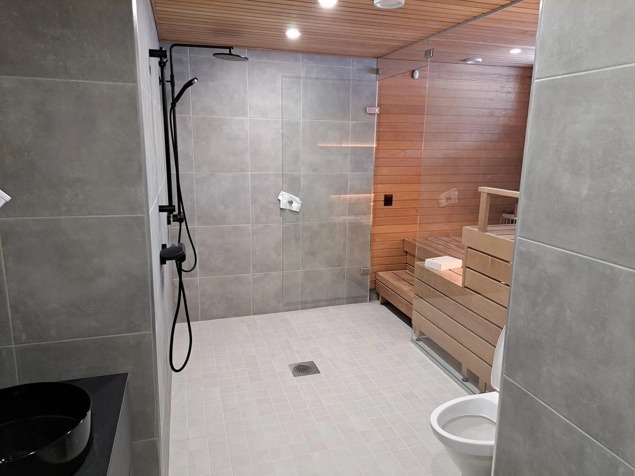 Kylpyhuoneessa on suihku, sauna ja wc, harmaata laattaa ja puupanelointia.