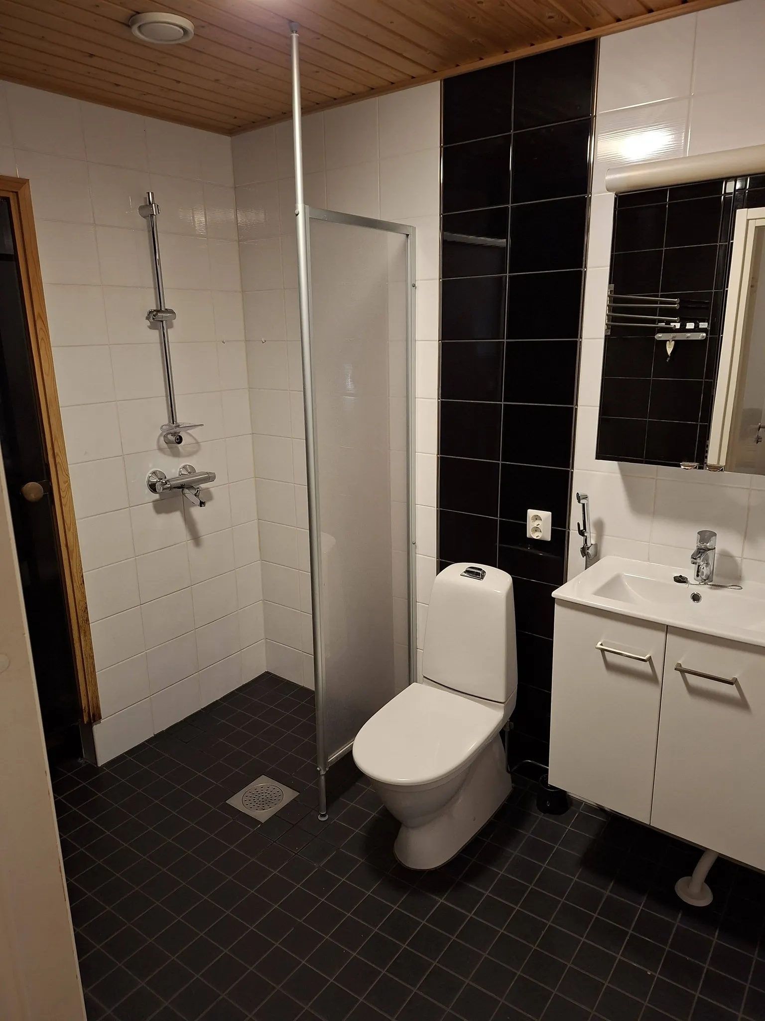 Kylpyhuoneessa on suihku, wc, pesuallas ja peilikaappi. Seinät ovat osittain valkoisia ja osittain mustia laattoja. Lattia on tummanharmaa.