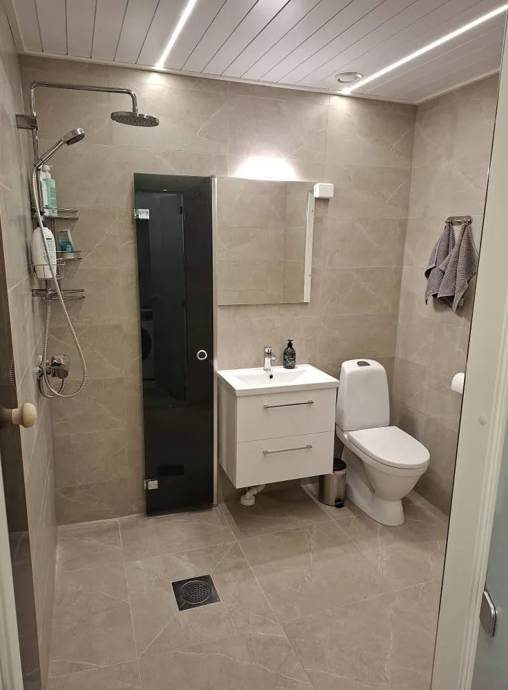 Kylpyhuoneessa on suihku, peili, pesuallas, wc ja tumma lasiovi. Seinät ja lattia ovat harmaata laattaa, katossa on valkoinen paneeli ja valaistus.