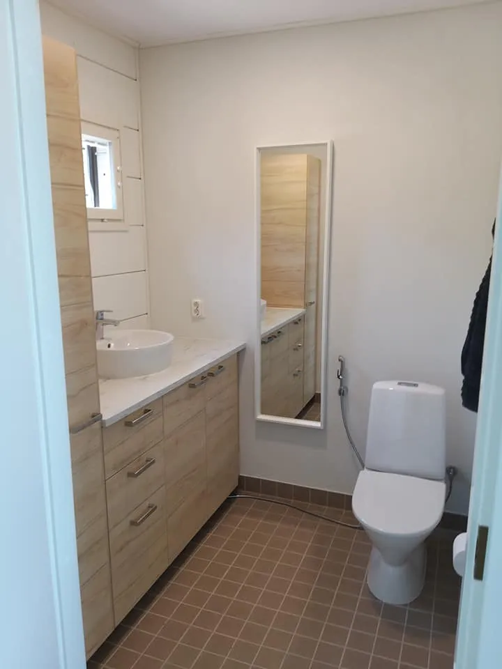 Valoisassa kylpyhuoneessa on puunsävyinen pesuallas, valkoinen posliiniallas, peili ja wc-istuin ruskealla laattalattialla.