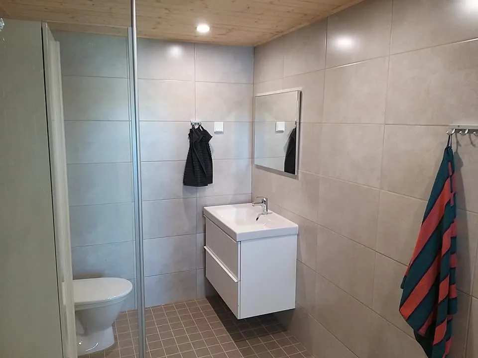 Kylpyhuoneessa on valkoiset kalusteet, harmaat laatat seinissä ja ruskea laattalattia. Suihkukaappi on lasinen ja katossa on puupanelointi.