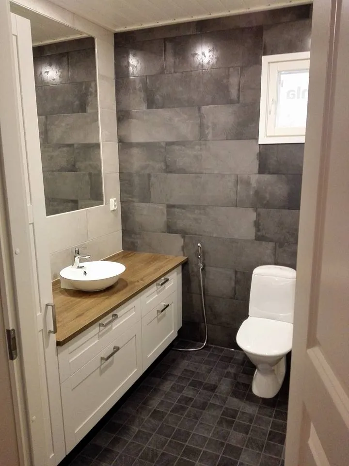 Kylpyhuoneessa on valkoinen pesuallas, puinen taso, harmaat seinälaatat, tummat lattialaatat ja valkoinen wc-istuin.