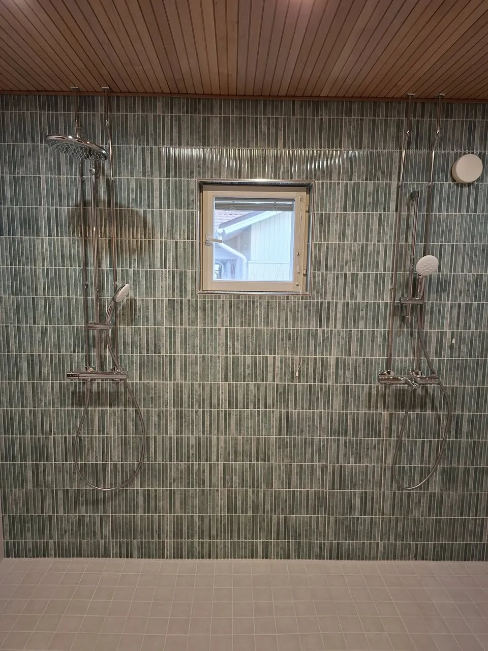 Kylpyhuoneen sisustus, jossa on kaksi suihkua, pieni ikkuna ja vihreät laatat sekä puinen katto.