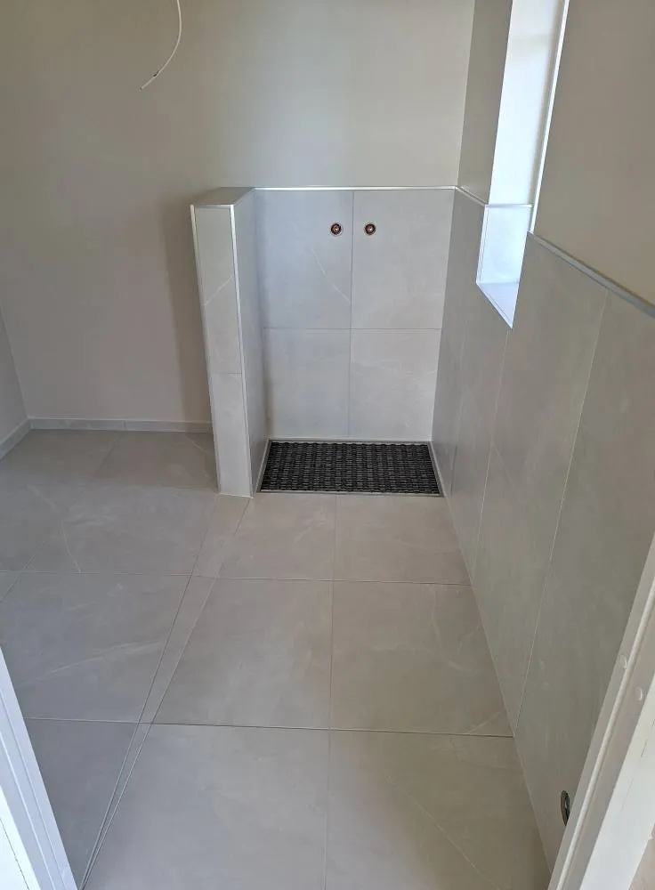 Vaaleanharmaa kylpyhuone, jossa on suuri suihkutila, harmaa ritilä ja valkoinen ikkuna.