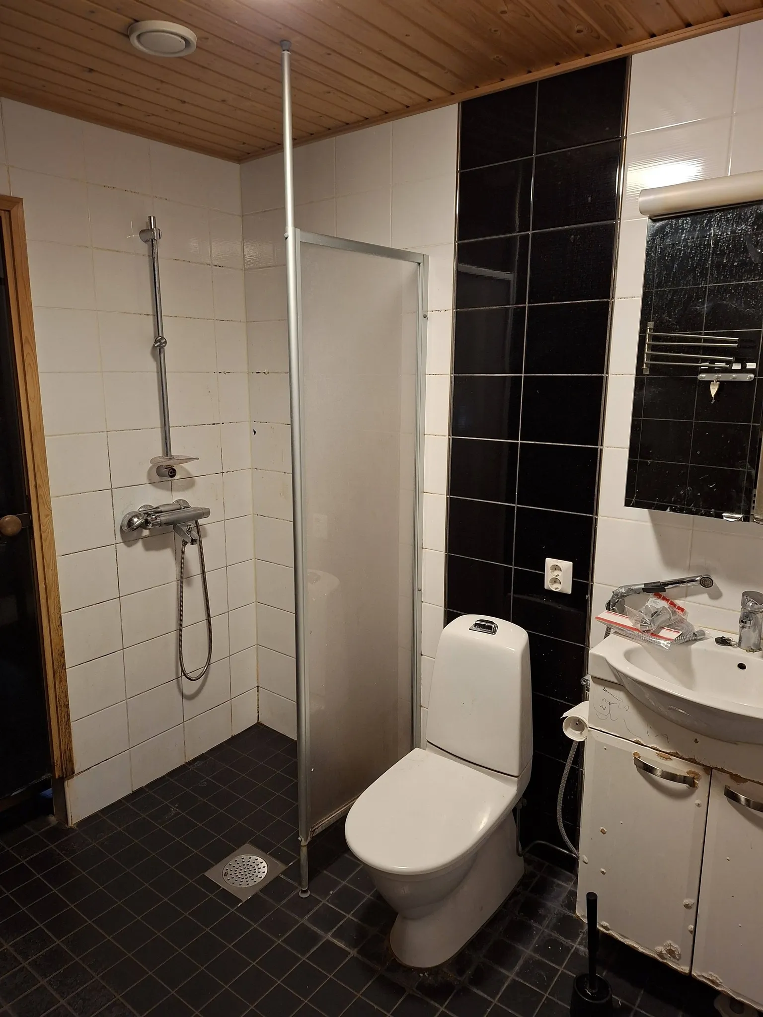 Kylpyhuoneessa on wc-istuin, suihku, pesuallas ja peili. Huoneessa on mustavalkoiset laatat ja puinen katto. Valo on himmeää.