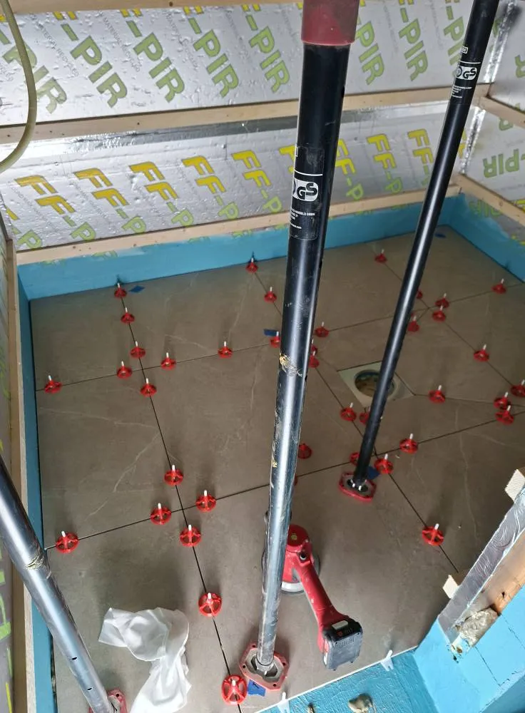 Valokuva näyttää kylpyhuoneen, jossa on juuri asennettu lattia. Lattialaatat ovat harmaita ja niiden välissä on punaisia kiiloja. Katossa on eristyslevyjä ja seinät on maalattu sinisiksi. Kolme metallista tukipylvästä on sijoitettu lattialle.