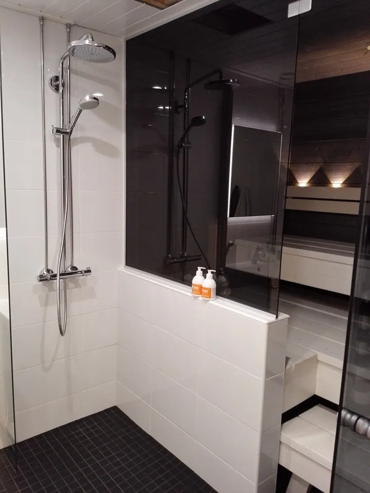 Kylpyhuoneen kuva, jossa on suihku, valkoiset laatat, tumma lattia ja kaksi oranssia pulloa.