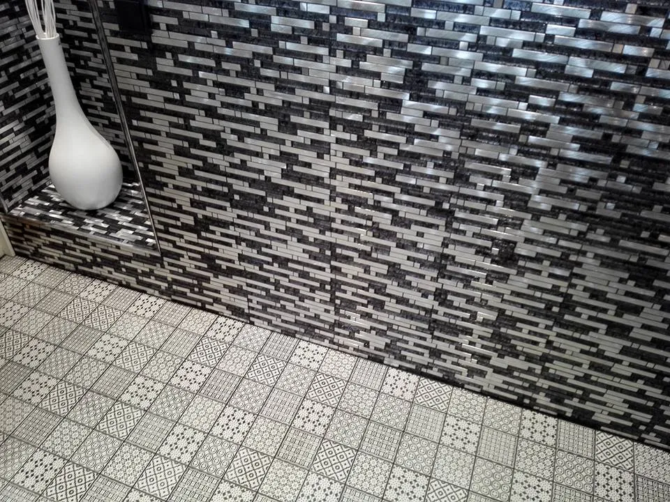 Kylpyhuoneen seinässä on mosaiikkilaattoja ja lattialla kuviollisia laattoja. Seinällä on valkoinen maljakko. Valoisa ja moderni sisustus.