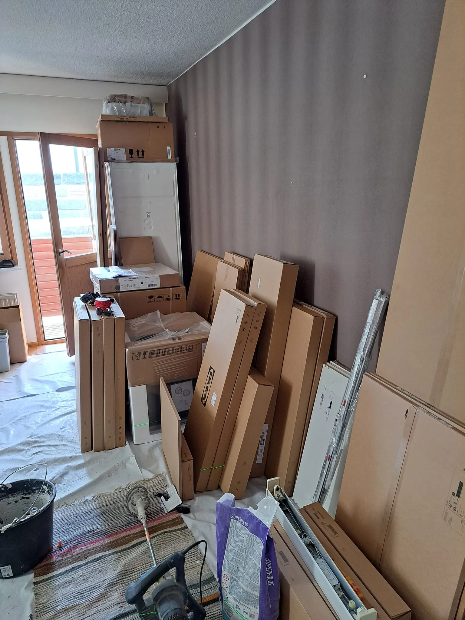 Huone täynnä pahvilaatikoita, remonttitarvikkeita ja valkoisia huonekaluja, valaistus on kirkas.