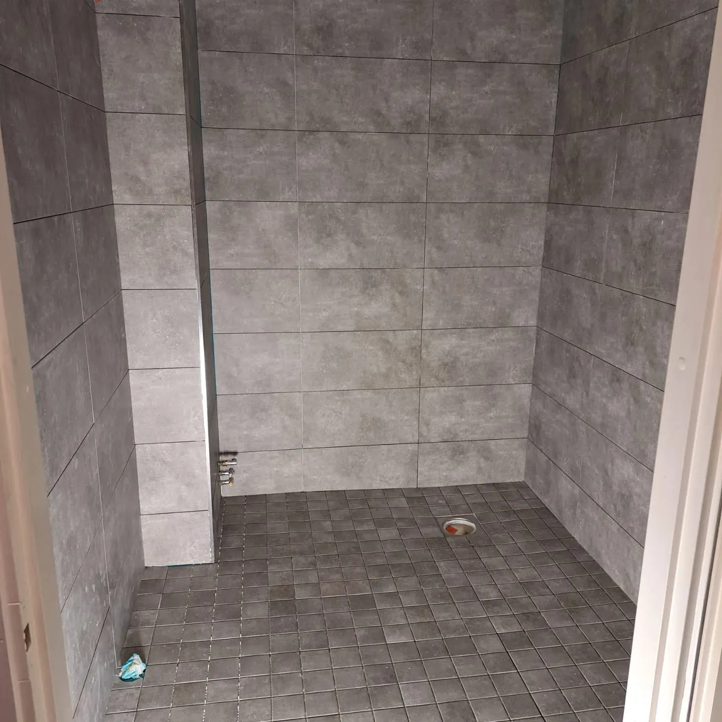 Harmaa laatoitettu suihkutila, jossa on lattiakaivo ja seinäkiinnikkeet, valaistus on himmeä.