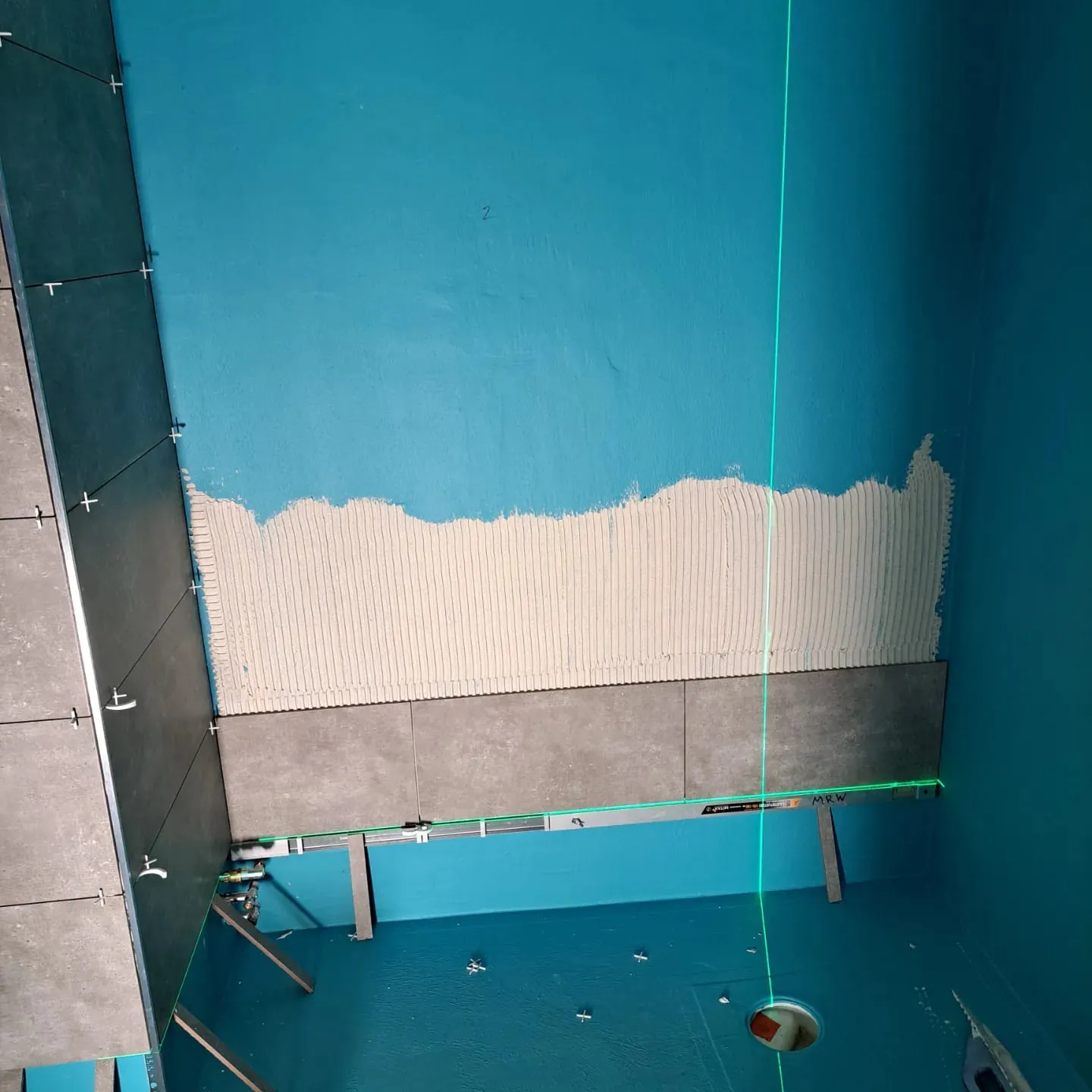 Valokuva rakennustyömaasta, jossa on siniset seinät, harmaat laatat, valkoinen laasti ja vihreä laserlinja.
