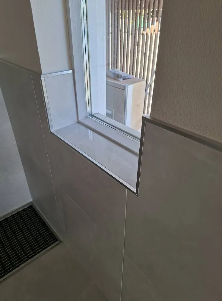 Kylpyhuoneen ikkuna, jossa on valkoiset laatat ja metalliset reunukset, sekä musta lattiaritilä. Kirkas valo.