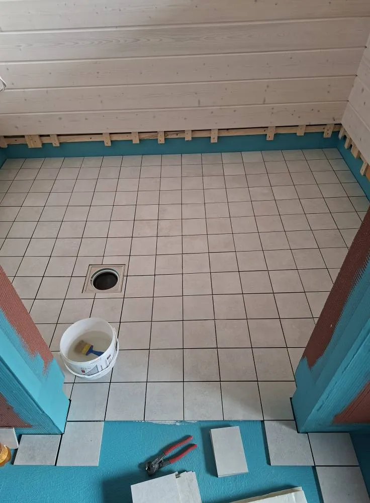 Huoneessa on vaaleat lattialaatat, valkoinen puukatto ja turkoosit seinät. Lattiassa on viemäri ja ämpäri, ja seinien alareunassa on puulistoja.