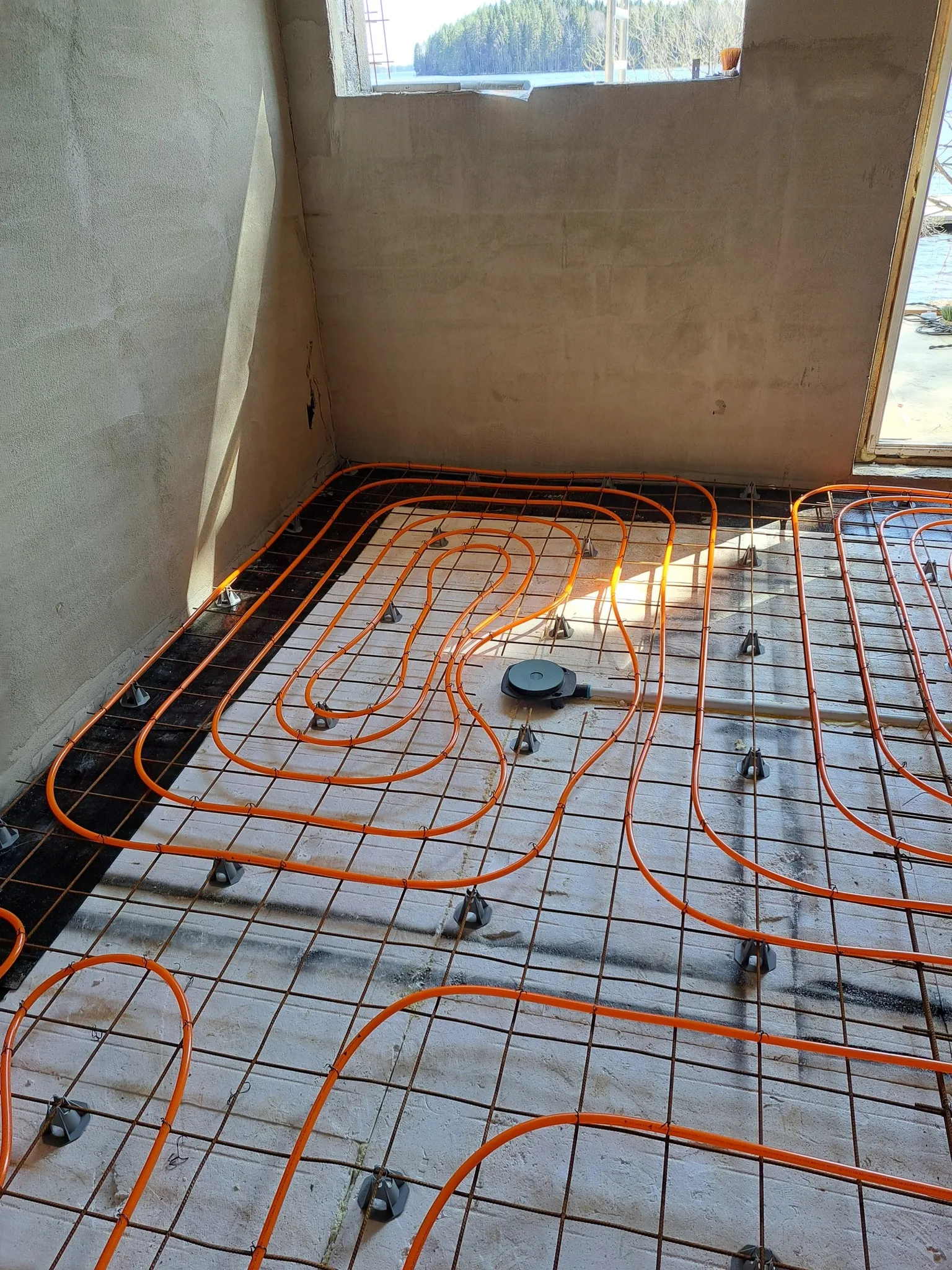 Huoneessa on lattialämmitysjärjestelmä, jossa on oranssit putket kiinnitettynä metalliverkkoon harmailla kiinnikkeillä, ja valo paistaa ikkunasta.