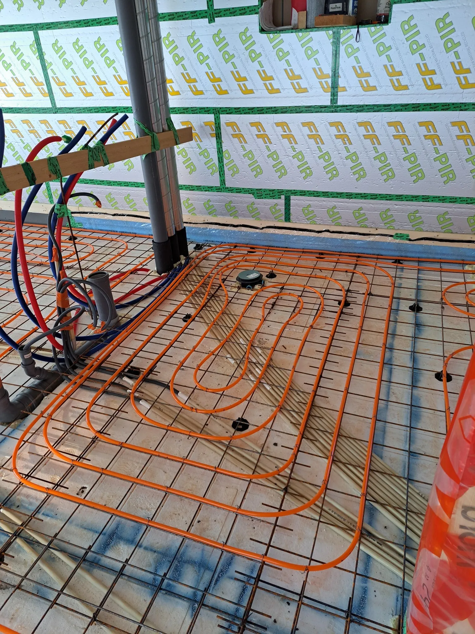 Lattialämmitysputket on asennettu metalliverkkoon rakennustyömaalla, taustalla eristyslevyt ja putket. Oranssit putket luovat geometrisen kuvion.