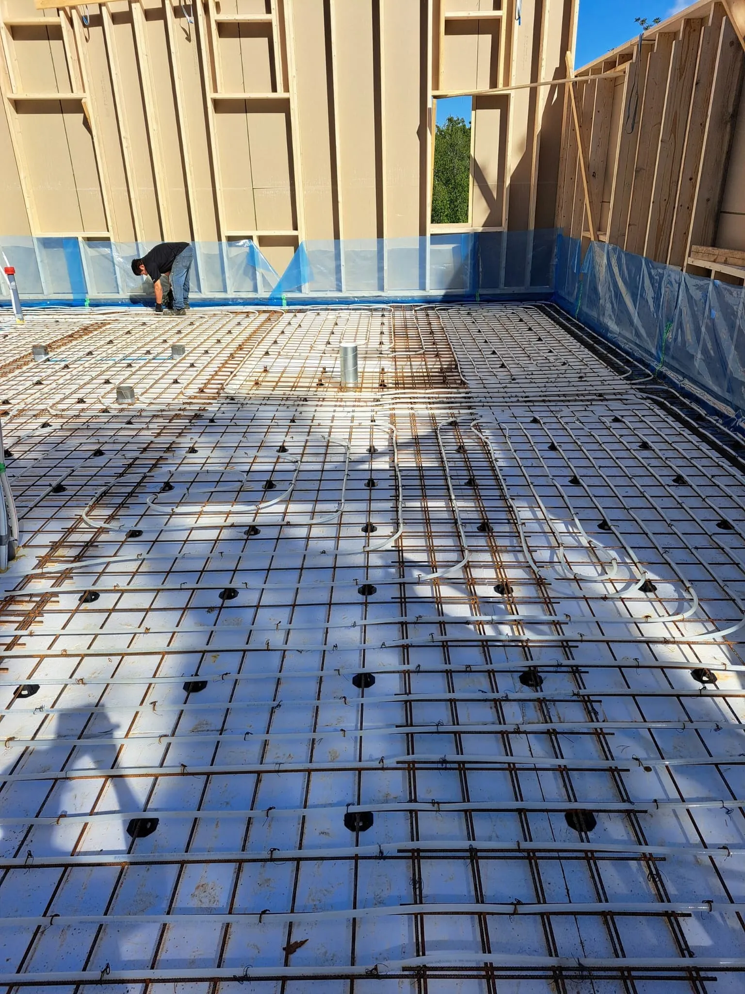 Rakennustyömaa, jossa lattialämmitysputket ja teräsverkko valkoisella eristeellä, mies työskentelee taustalla, valoisa sää.
