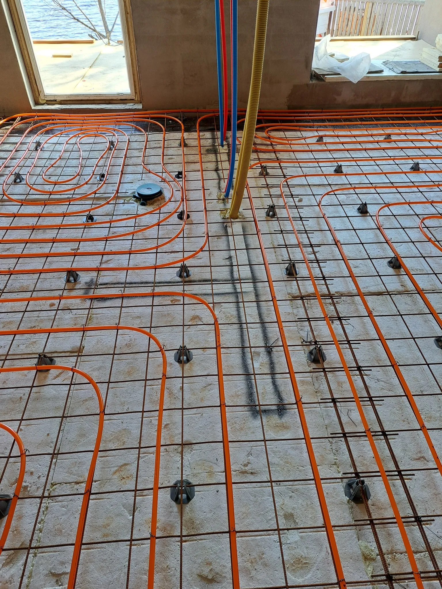 Lattialämmitysputket on asennettu raudoitusverkkoon, jossa on muovikiinnikkeet. Putket ovat oranssit, ja taustalla näkyy ikkuna ja rakennustarvikkeita.